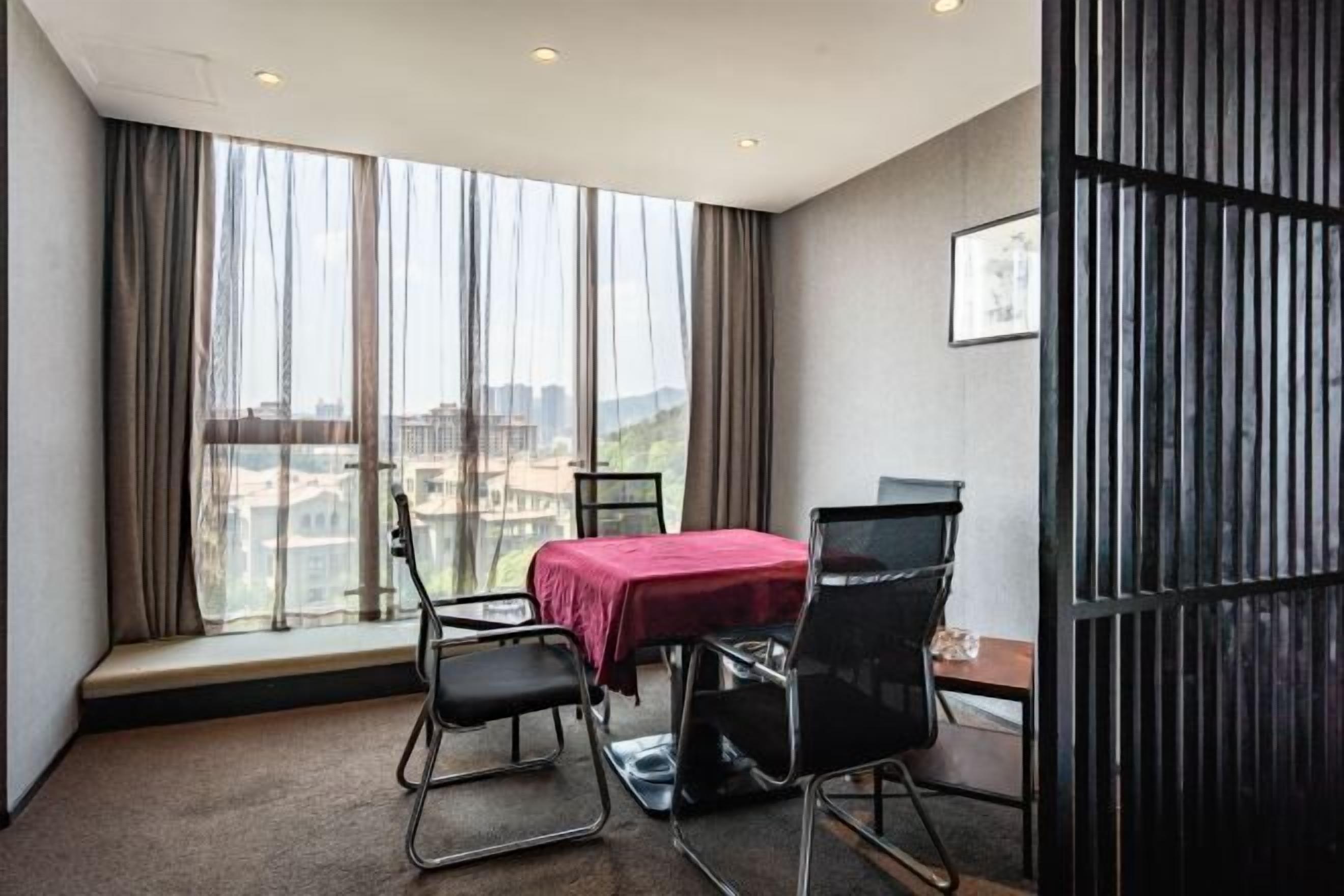 Une chambre d’hôtel avec une grande fenêtre, une table recouverte d’une nappe rouge et deux chaises noires.