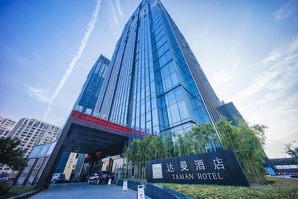 Un hôtel moderne avec une façade en verre remarquable, une enseigne indiquant qu’il s’agit de l’hôtel Taman, et un ciel dégagé en arrière-plan.