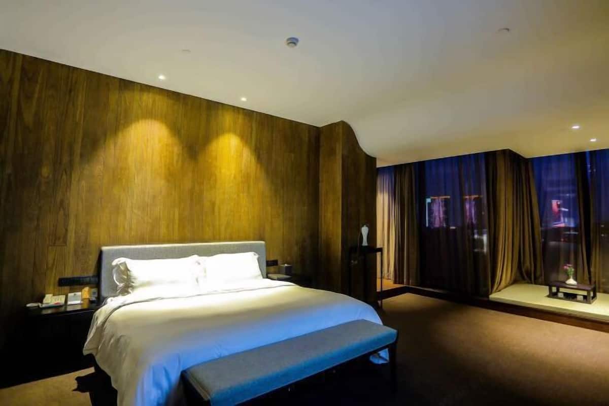 A modern hotel room with a large bed, a bedside table, a nightstand, a small table with a vase, and a window with curtains.