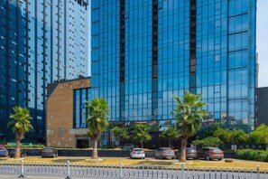 A modern high-rise building with reflective glass windows, palm trees in front, and cars parked along the street. - Tonglu Daman Hotel (Fuchunjiang , Tonglu City Center, Hangzhou) (Tonglu)