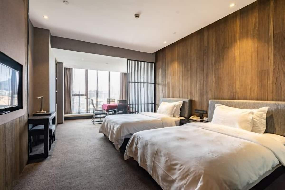 A hotel room with two beds, a desk, a chair, and a large window offering a city view.