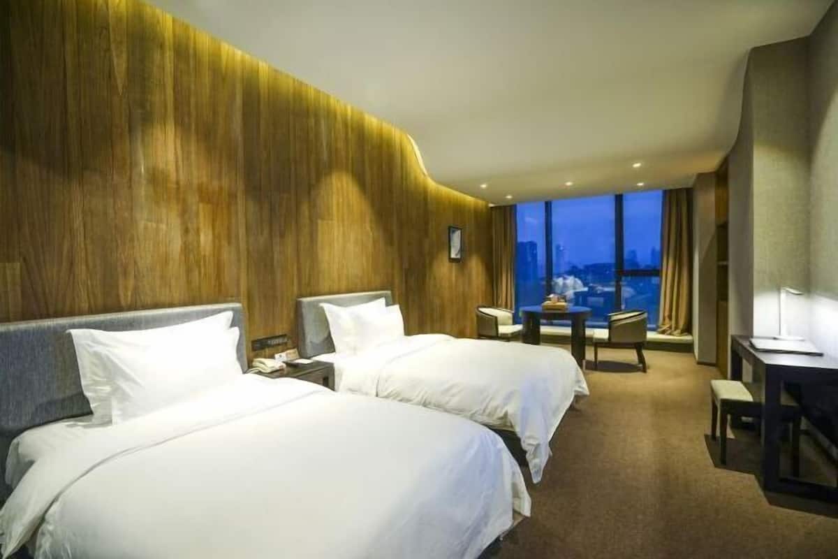 A hotel room with two beds, a desk, a chair, a small table, and a view of the cityscape.