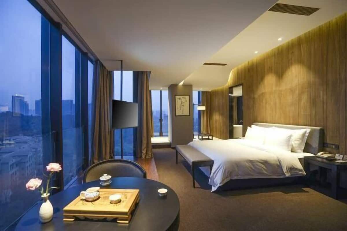 A modern hotel room with a large window, a bed, a bedside table, and a small dining table with a vase of flowers.