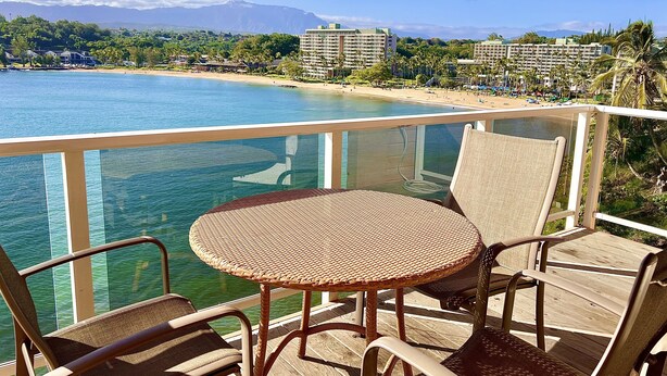 A balcony with wicker chairs and a table overlooking a beach with buildings in the background. - Oceanfront home on one of the most popular beaches in Kauai! (Lihue)