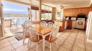 A kitchen with wooden cabinets, a dining area with a table and chairs, and a view of a pool and mountains. - Oceanfront home on one of the most popular beaches in Kauai! (Lihue)