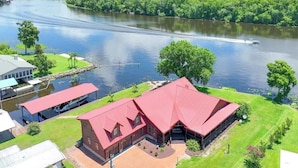 A large, red-roofed building with a spacious lawn and a dock extending into a river.