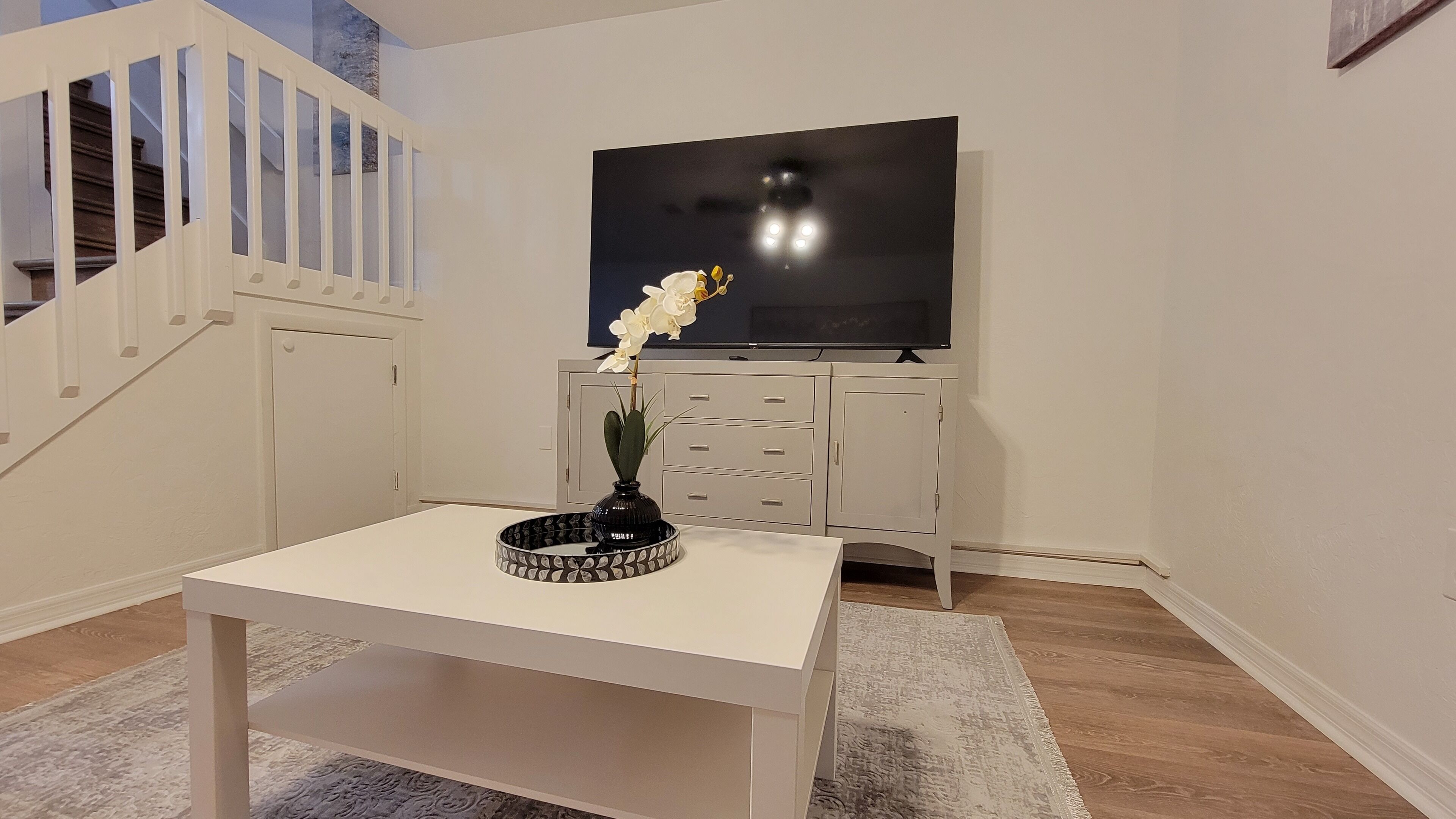 A modern living room with a white table, a black vase with white flowers, a TV on the wall, and a staircase.