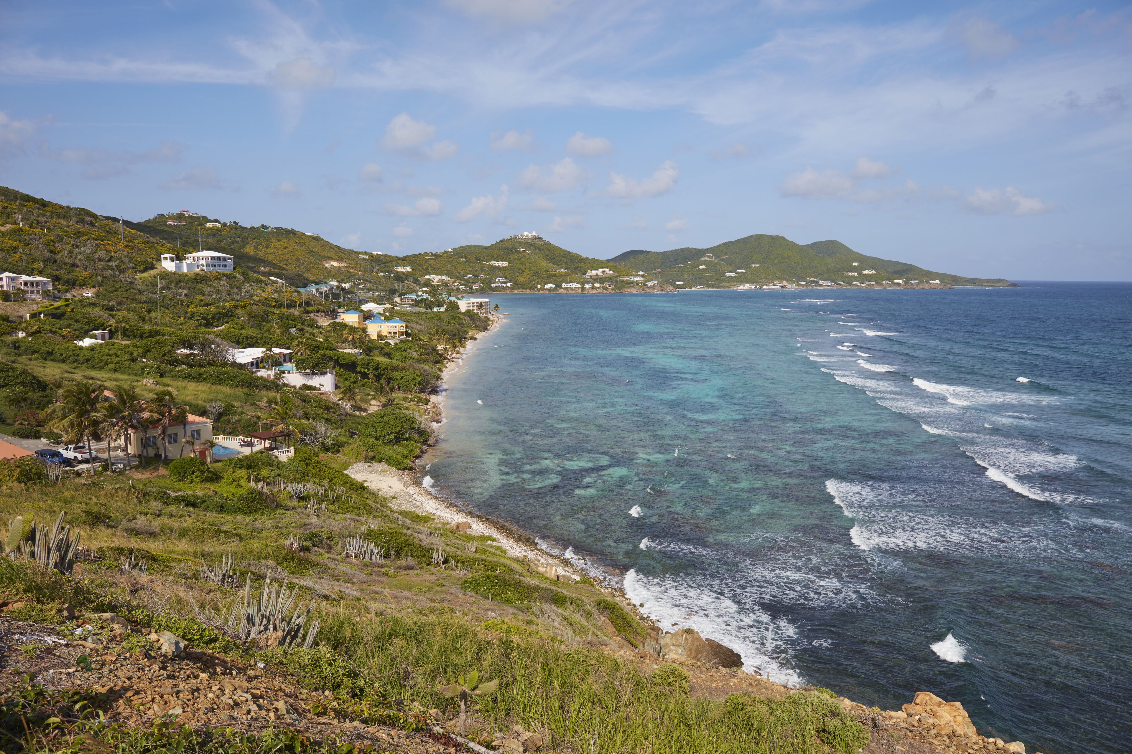 A coastal landscape with a clear view of the ocean, a beach, and a hillside with buildings.