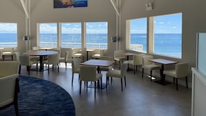 A spacious dining area with large windows offering ocean views, white chairs, and tables.