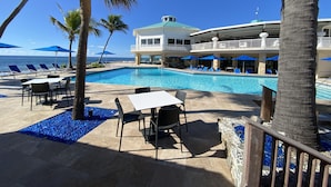 A pool area with lounge chairs and umbrellas, a pool with clear blue water, and a building with a balcony overlooking the pool.