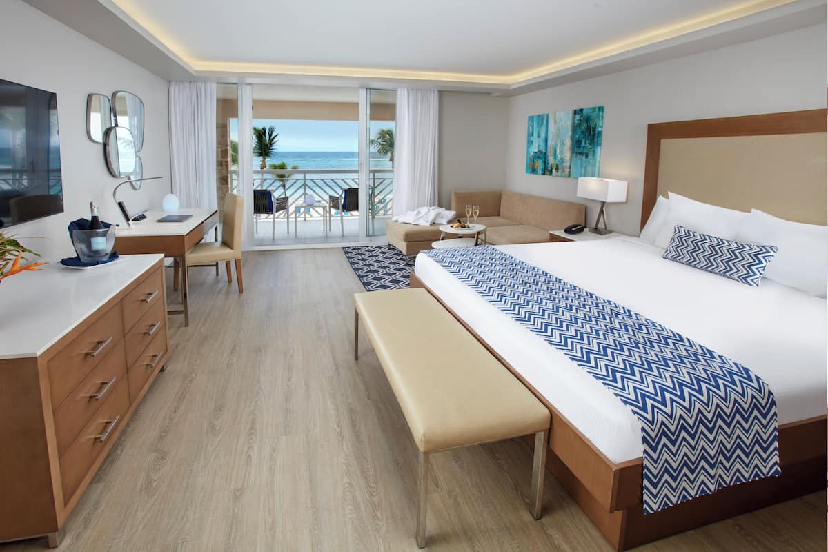 Een hotelkamer met een groot bed, een bureau, een stoel, een bank, een televisie en een balkon met uitzicht op de oceaan.