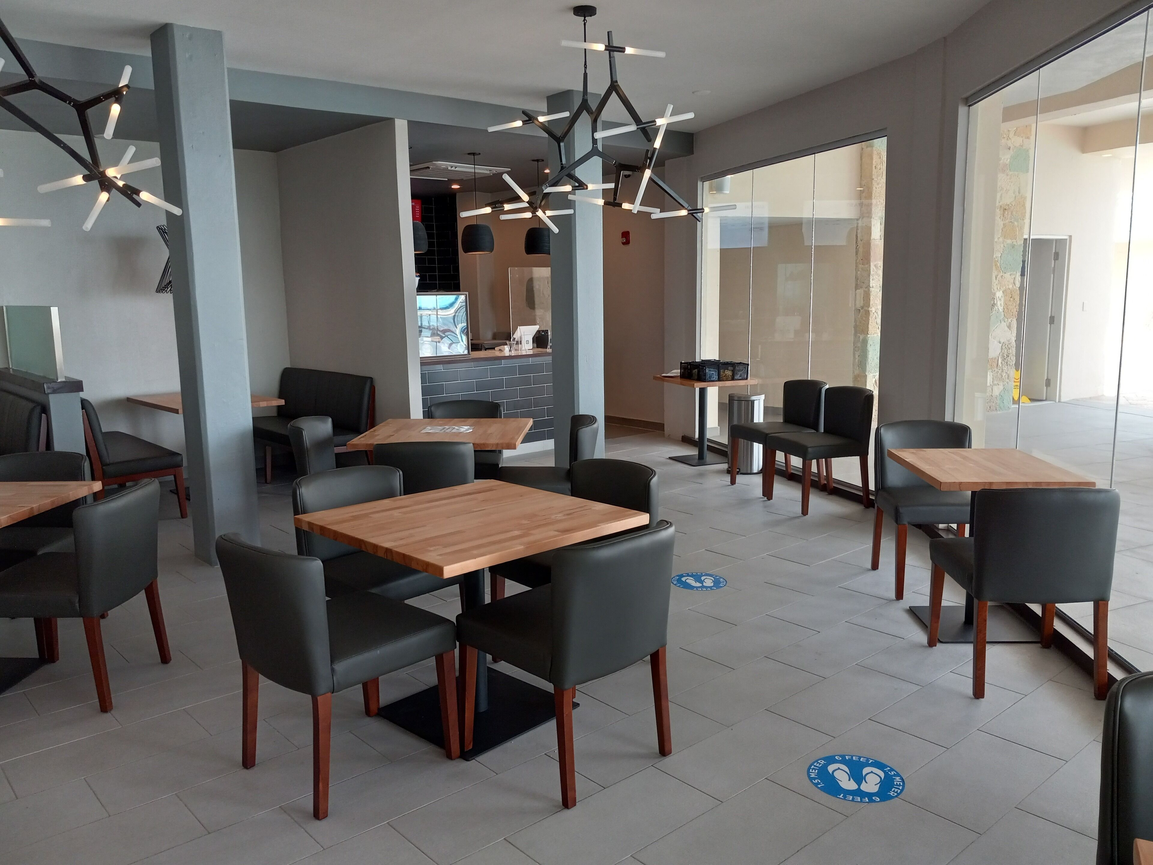 Ein modernes Restaurant mit Holztischen und schwarzen Stühlen, einem Küchenbereich im Hintergrund und einem großen Fenster, das für natürliches Licht sorgt.