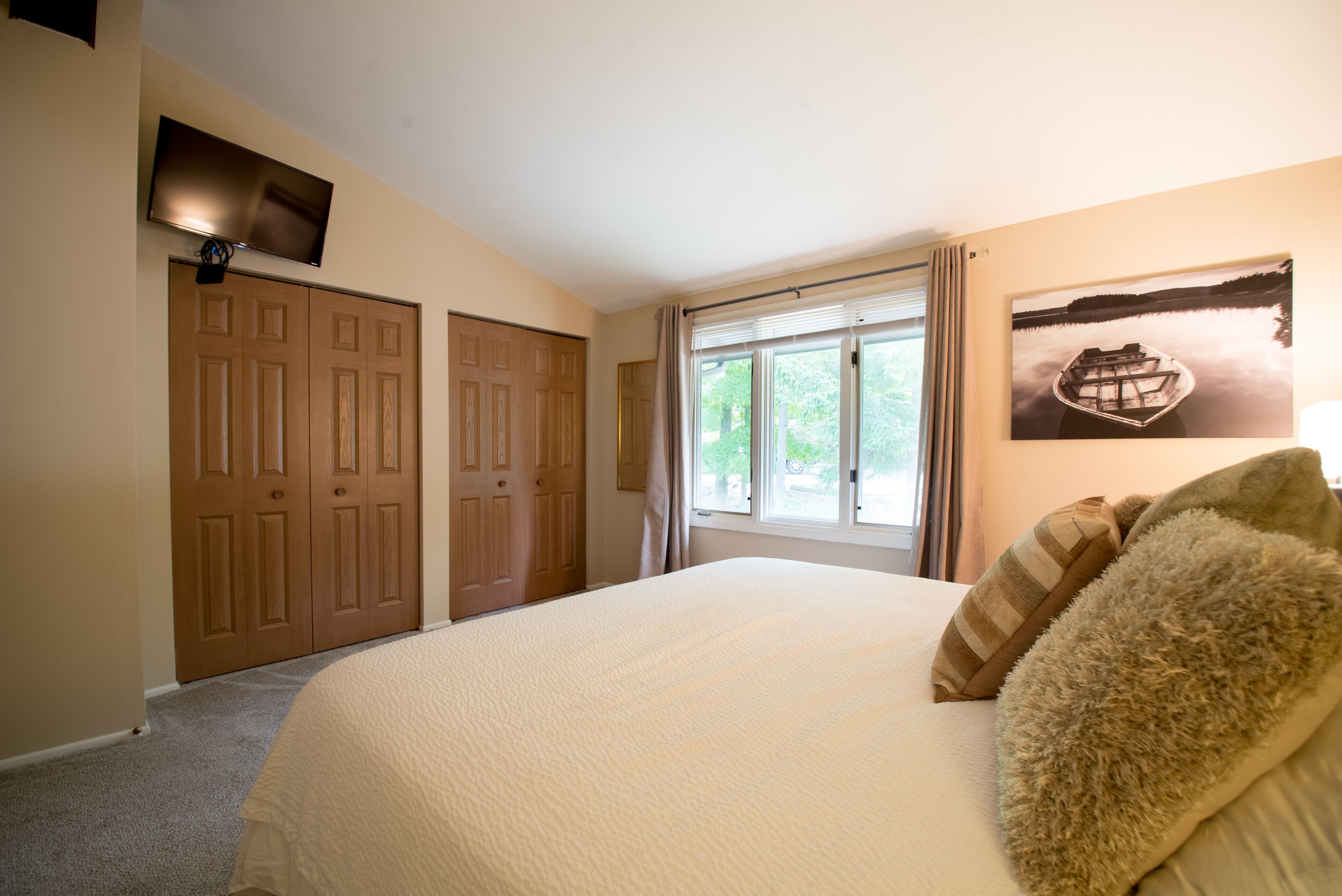 A bedroom with a large bed, a flat-screen TV mounted above a wooden door, a window with curtains, and a framed picture on the wall.
