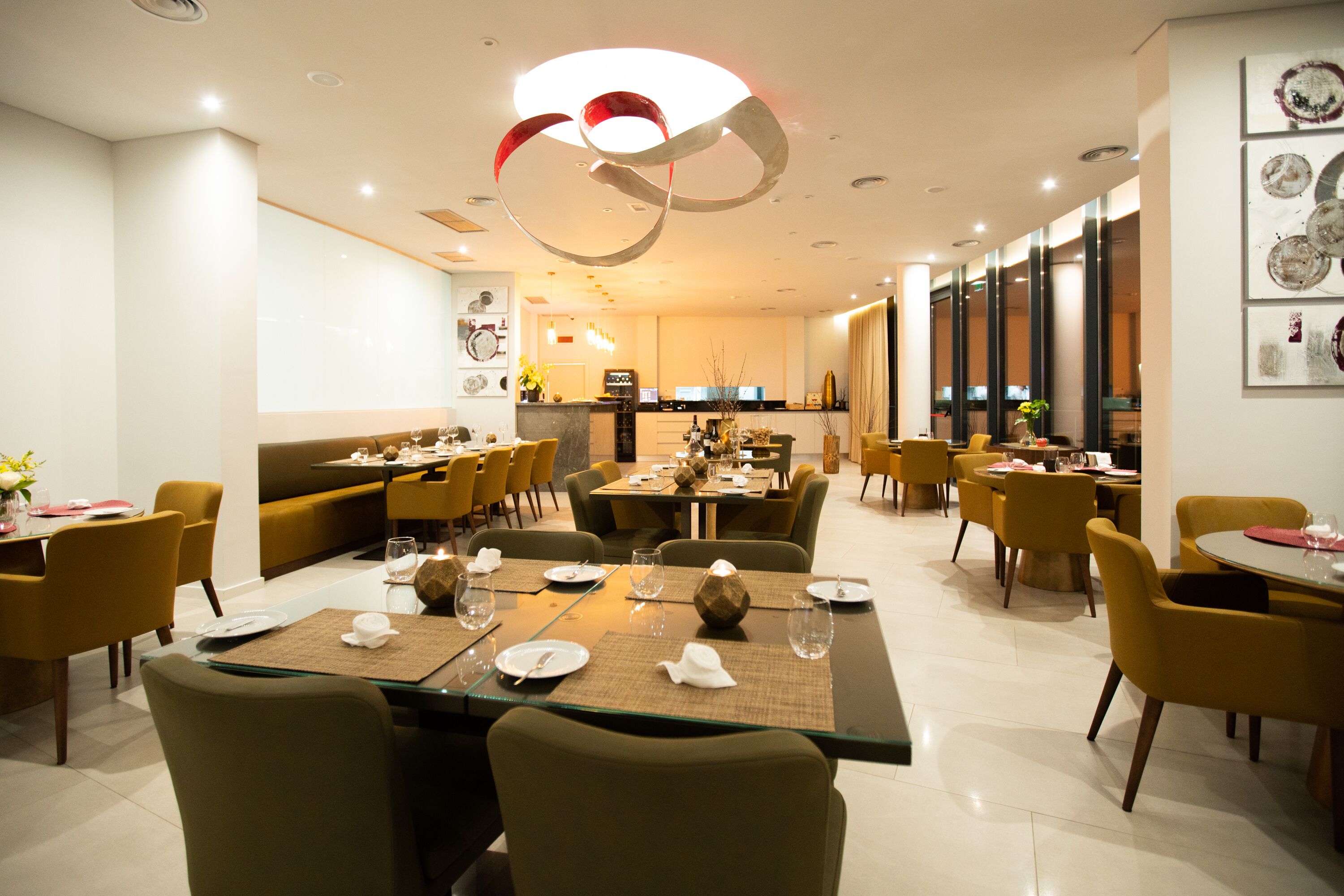 Uma sala de jantar moderna com mesas preparadas para o serviço, uma cozinha ao fundo e um ambiente claro e bem iluminado.