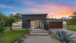 Une maison moderne à deux étages, dotée d’un garage, d’une entrée avec une porte vitrée et d’un jardin bien entretenu où poussent différentes espèces de plantes.