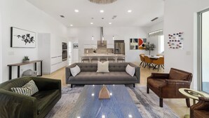 Un salon moderne avec un canapé, une table basse et des fauteuils, une cuisine en arrière-plan et des œuvres d’art accrochées aux murs.