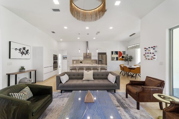 A modern living room with a sofa, coffee table, and chairs, a kitchen in the background, and artwork on the walls.