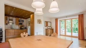 Un vaste espace de salle à manger avec une grande table en bois, une cuisine équipée d’appareils modernes et de grandes fenêtres offrant une vue sur l’extérieur.
