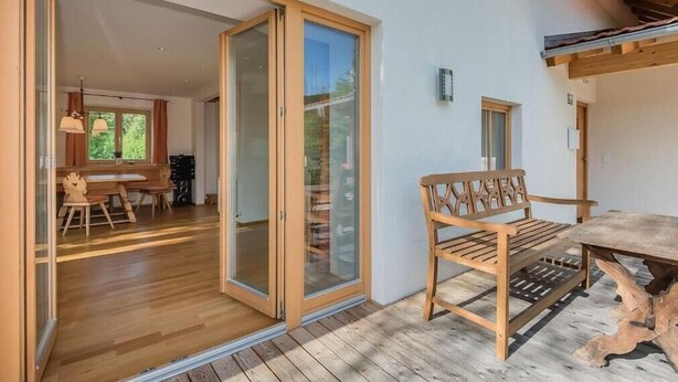 Une table et un banc en bois sont installés sur une terrasse en bois. Une porte-fenêtre coulissante donne accès à un espace intérieur où se trouvent une table et des chaises en bois.