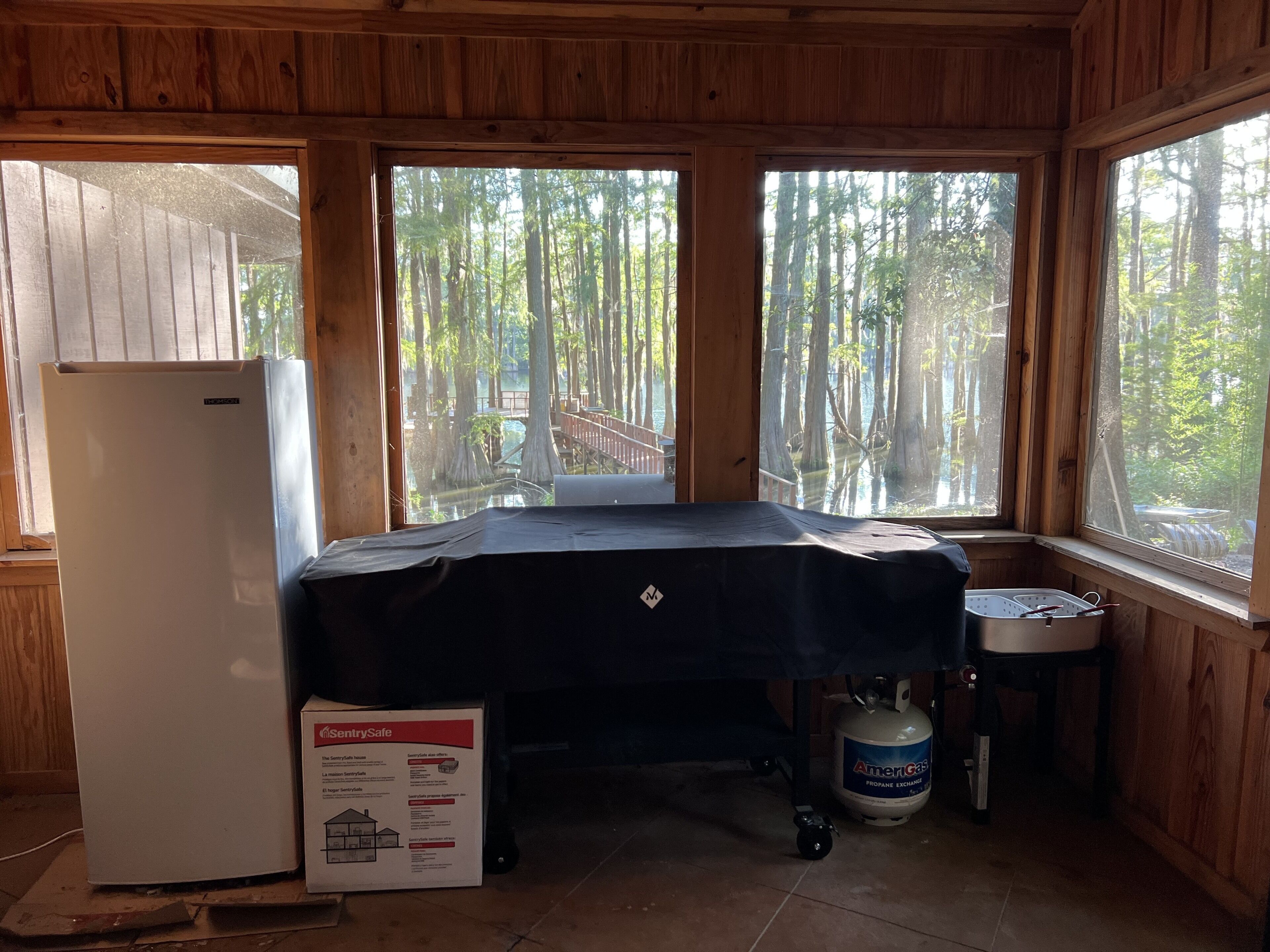 A cabin interior with a table, a refrigerator, a propane tank, and large windows offering a forest view.