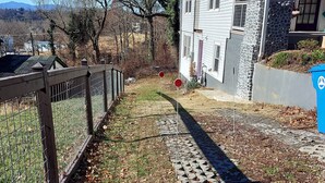 Un chemin pavé, bordé d’une clôture d’un côté et d’un bâtiment à la façade en pierre et au porche de l’autre.