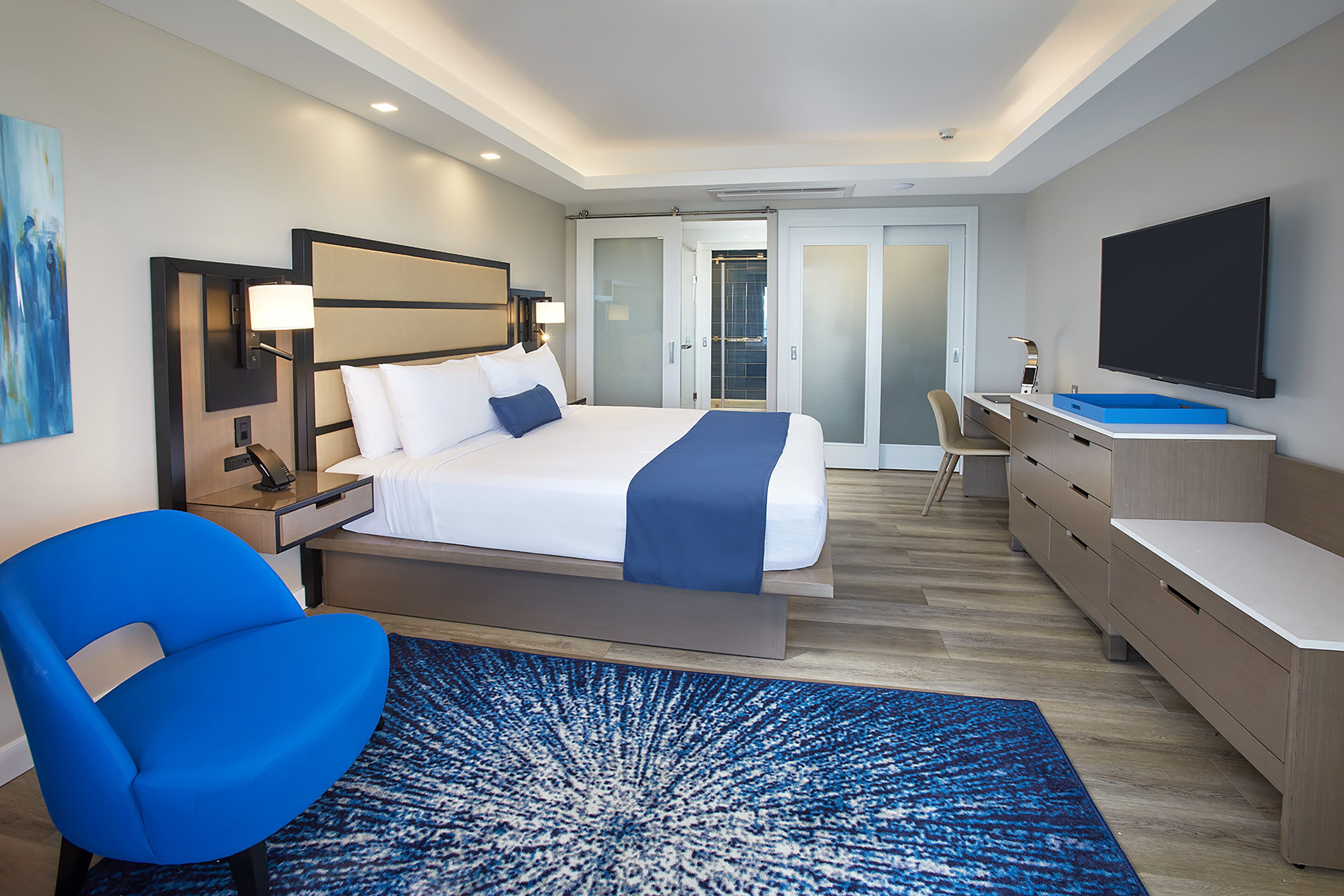 Ein modernes Hotelzimmer mit einem großen Bett, einem Schreibtisch, einem Stuhl, einem Fernseher und einem durch eine Glastür einsehbarer Badezimmer.