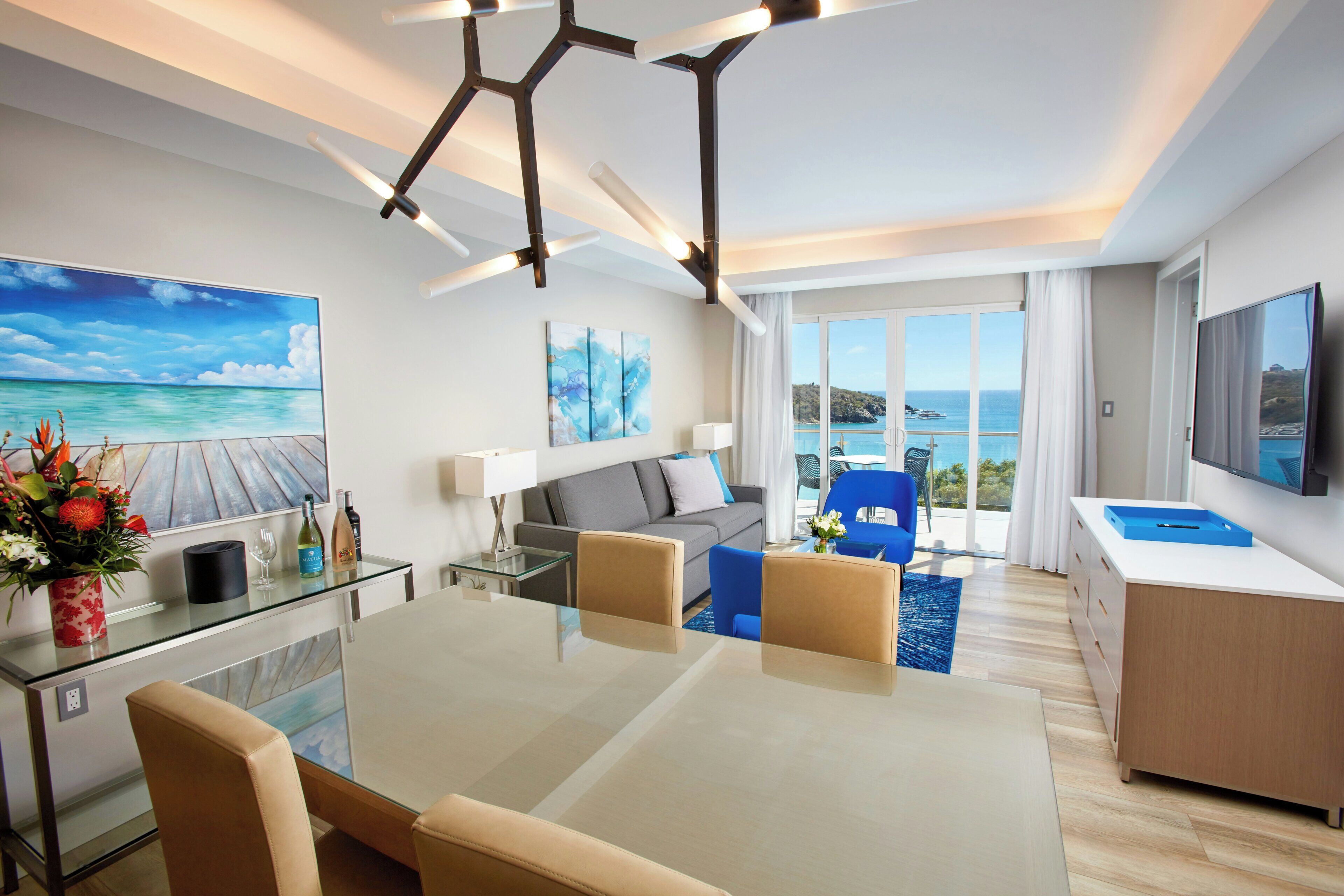 A modern hotel room with a dining area, a sofa, a TV, and a view of the sea.