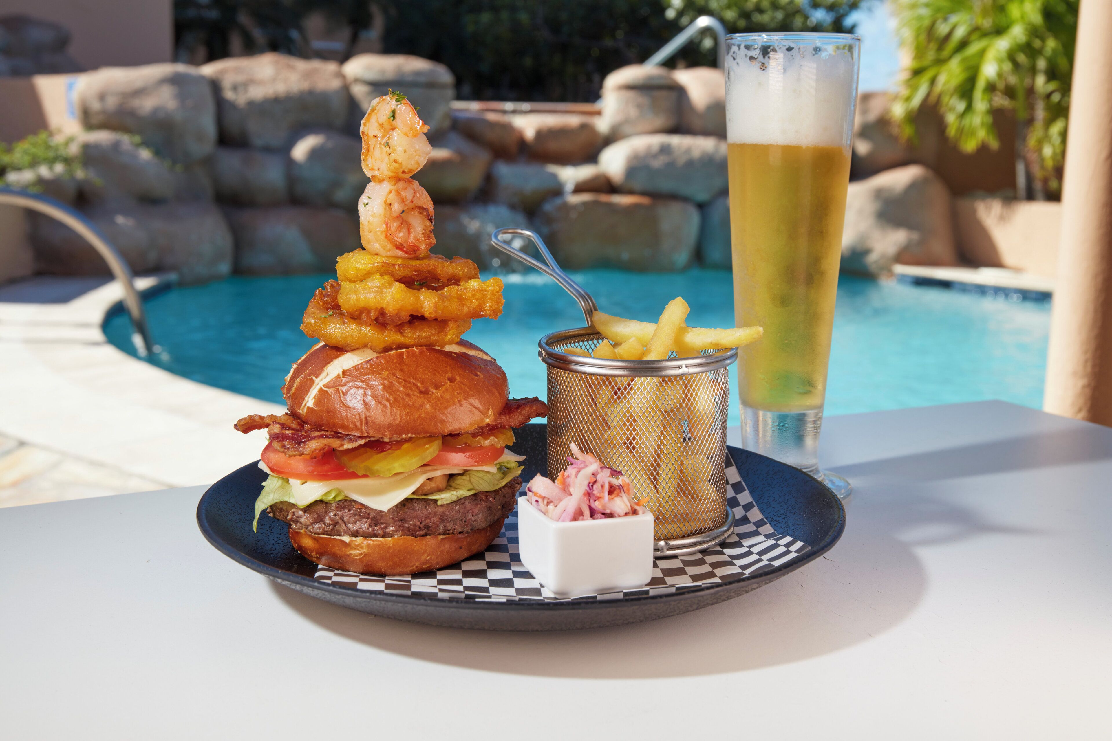 A meal with a burger, onion rings, and a drink by a poolside.