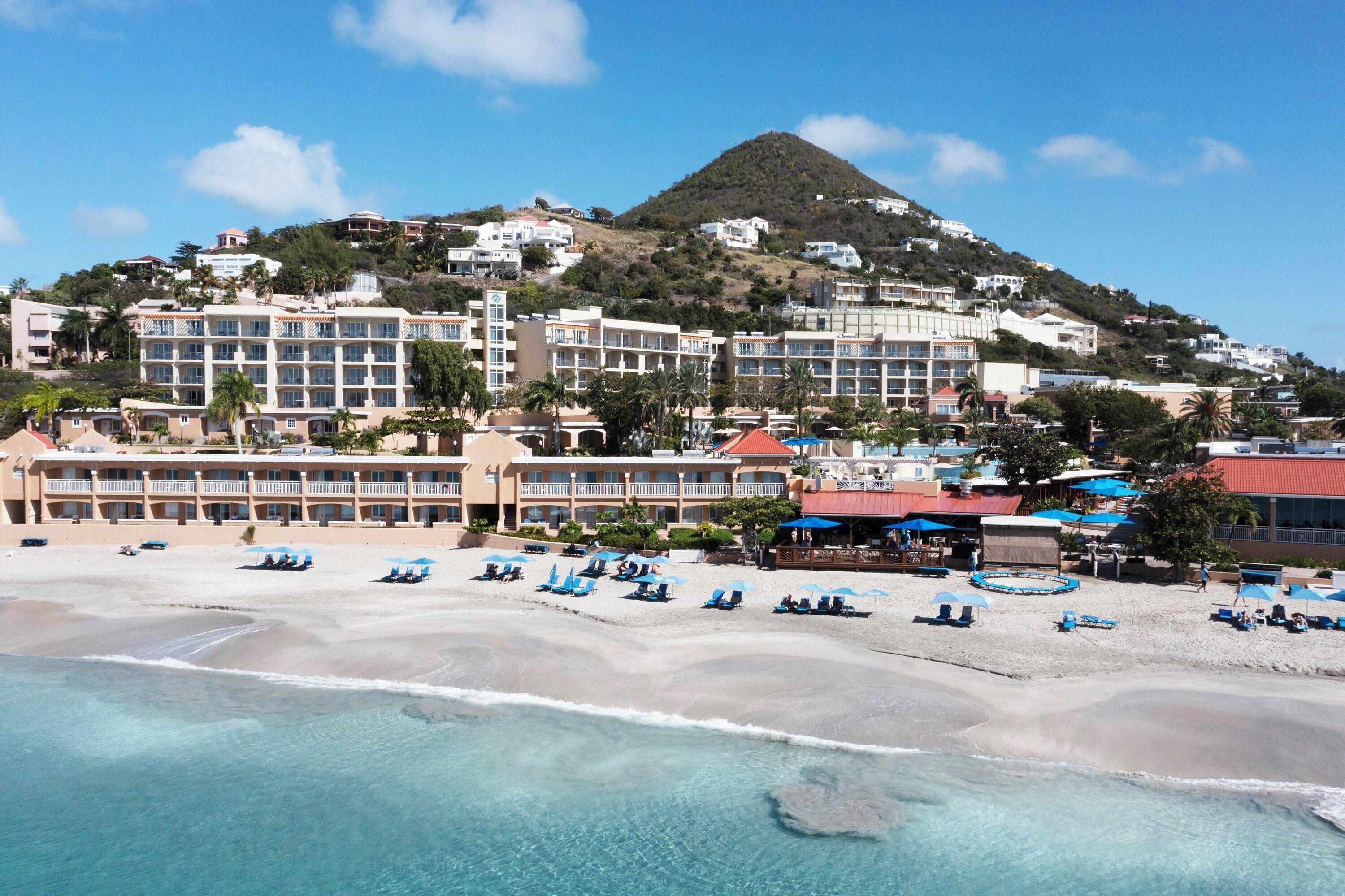 Un hotel frente a la playa con varias edificaciones, una playa con tumbonas y una colina al fondo.