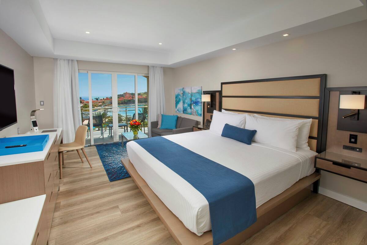 A modern hotel room with a large bed, a desk with a computer, a TV, and a view of a pool and buildings.