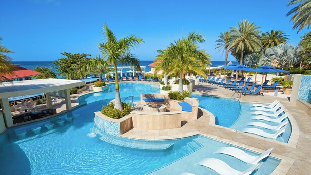 Ein Poolbereich mit Liegestühlen, Palmen und Blick aufs Meer.