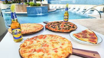 A poolside table with a variety of pizzas, a bottle of Carib beer, and a pool in the background.