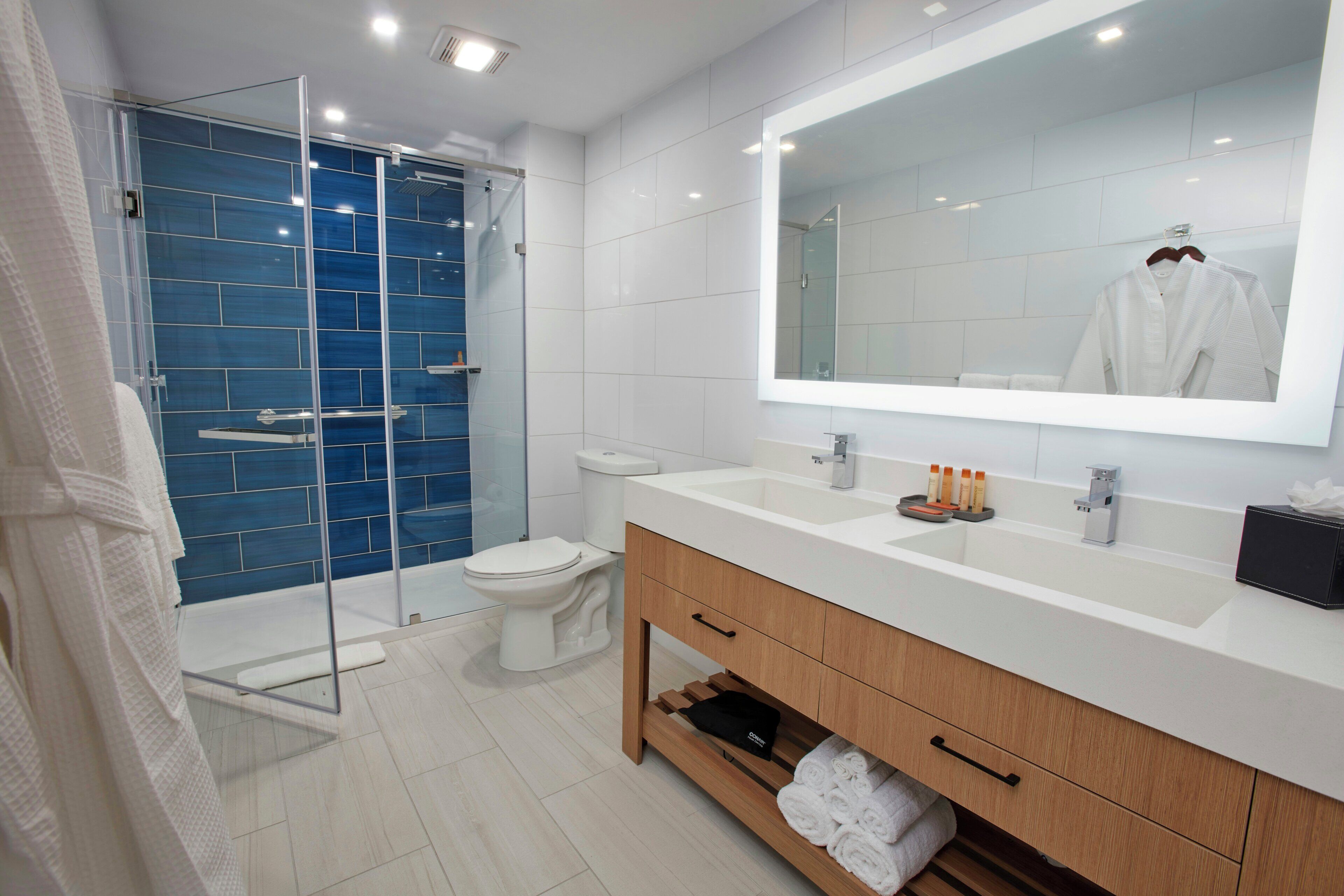 Un baño moderno con cabina de ducha de vidrio, inodoro blanco, espejo grande y tocador de madera con lavamanos.