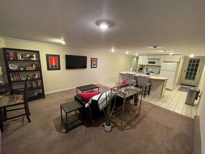 Un espace de vie compact comprenant un canapé, une table basse, une bibliothèque, un téléviseur à écran plat et un coin cuisine.