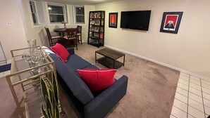 Una sala de estar con un sofá azul oscuro, cojines rojos, una mesa de centro de cristal, una estantería, un televisor de pantalla plana y una zona de comedor con una mesa y sillas.