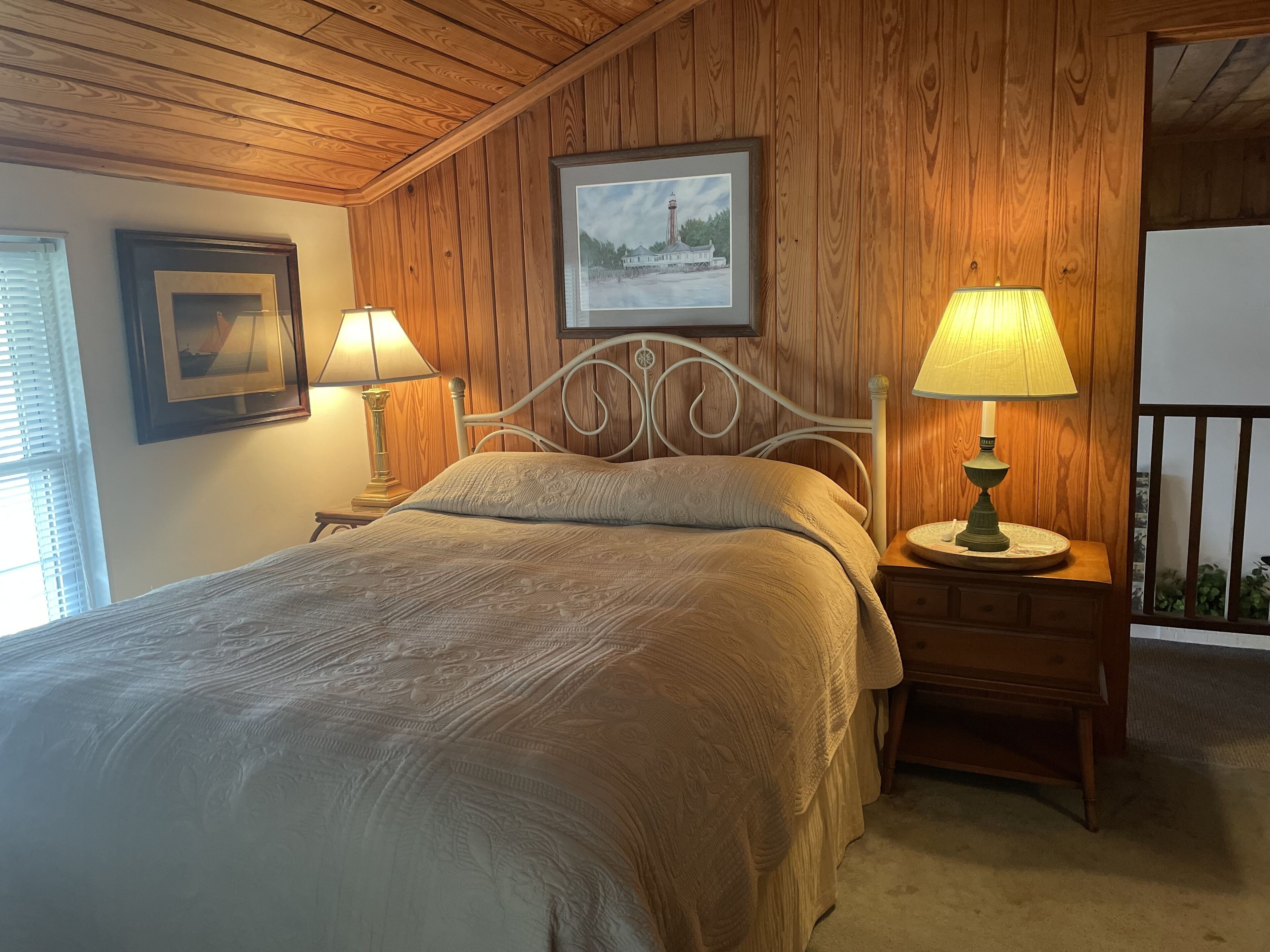 A bedroom with a wooden ceiling, a bed with a beige quilt, two bedside lamps, a framed picture on the wall, and a window with blinds.
