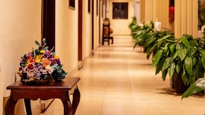 Un pasillo con vigas de madera en el techo, un arreglo floral sobre una mesa y plantas en macetas a lo largo de las paredes.
