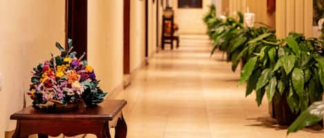 A corridor with wooden beams on the ceiling, a floral arrangement on a table, and potted plants along the walls.