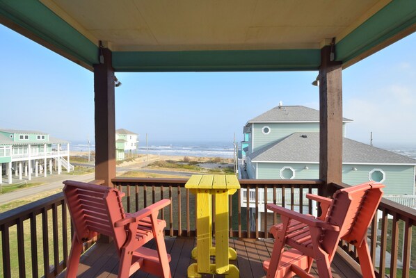Eine Holzterrasse mit roten Stühlen und einem gelben Tisch, mit Blick auf einen Strand und Gebäude.