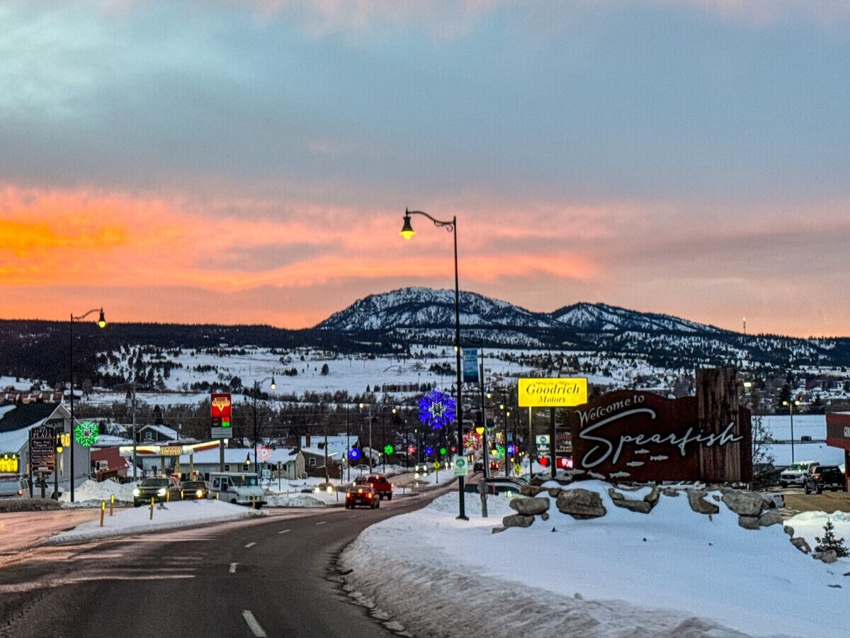 Un pueblo cubierto de nieve con una montaña al fondo, una estación de servicio y un letrero de bienvenida a Spearfish.