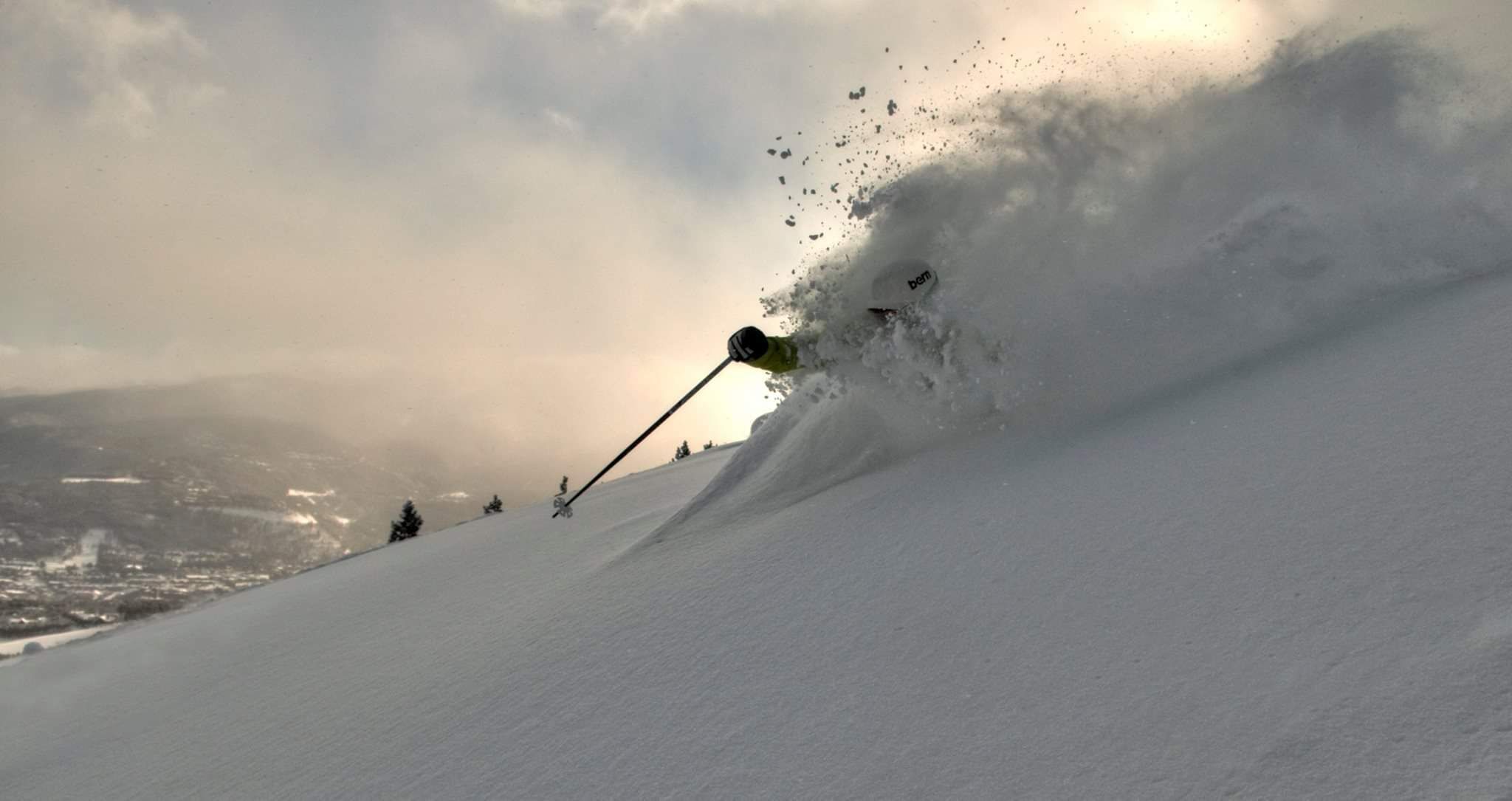 Eine Person fährt mit Skiern einen verschneiten Hang hinunter, einen Stock in der Hand, umgeben von Schnee und einem bewölkten Himmel.
