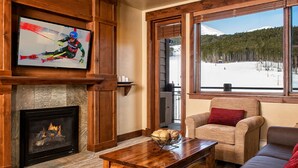 Un salón acogedor con chimenea, un televisor montado en una pared de madera, un sofá cómodo y un amplio ventanal con vistas a un paisaje nevado.