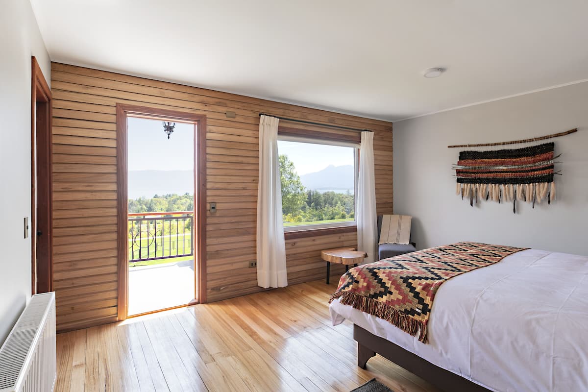 Un dormitorio con piso de madera, una cama con manta estampada, ventana con cortinas y vista a zonas verdes y montañas.