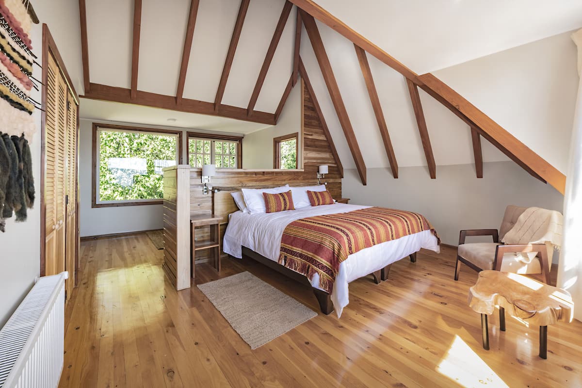 Un dormitorio con techo inclinado, vigas de madera, una cama con manta a rayas, una silla y una ventana con vistas al verdor.