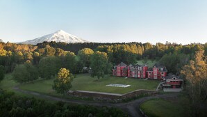 A large red building with multiple sections, surrounded by a green landscape and a snow-capped mountain in the background. - Das Dorf (Pucón)