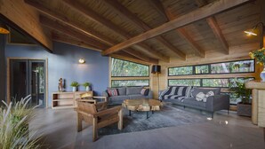 A spacious living area with a large gray sofa, wooden coffee table, and a wooden bench. There are two windows providing natural light and a view of the outdoors. - Das Dorf (Pucón)