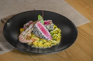 Un plato negro con un guiso de mariscos, incluyendo camarones y una porción de pescado, sobre una mesa de madera.
