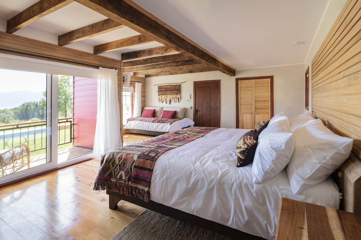 Un dormitorio espacioso con una cama grande, vigas de madera, un balcón con vistas y una puerta corredera que da a otra habitación.