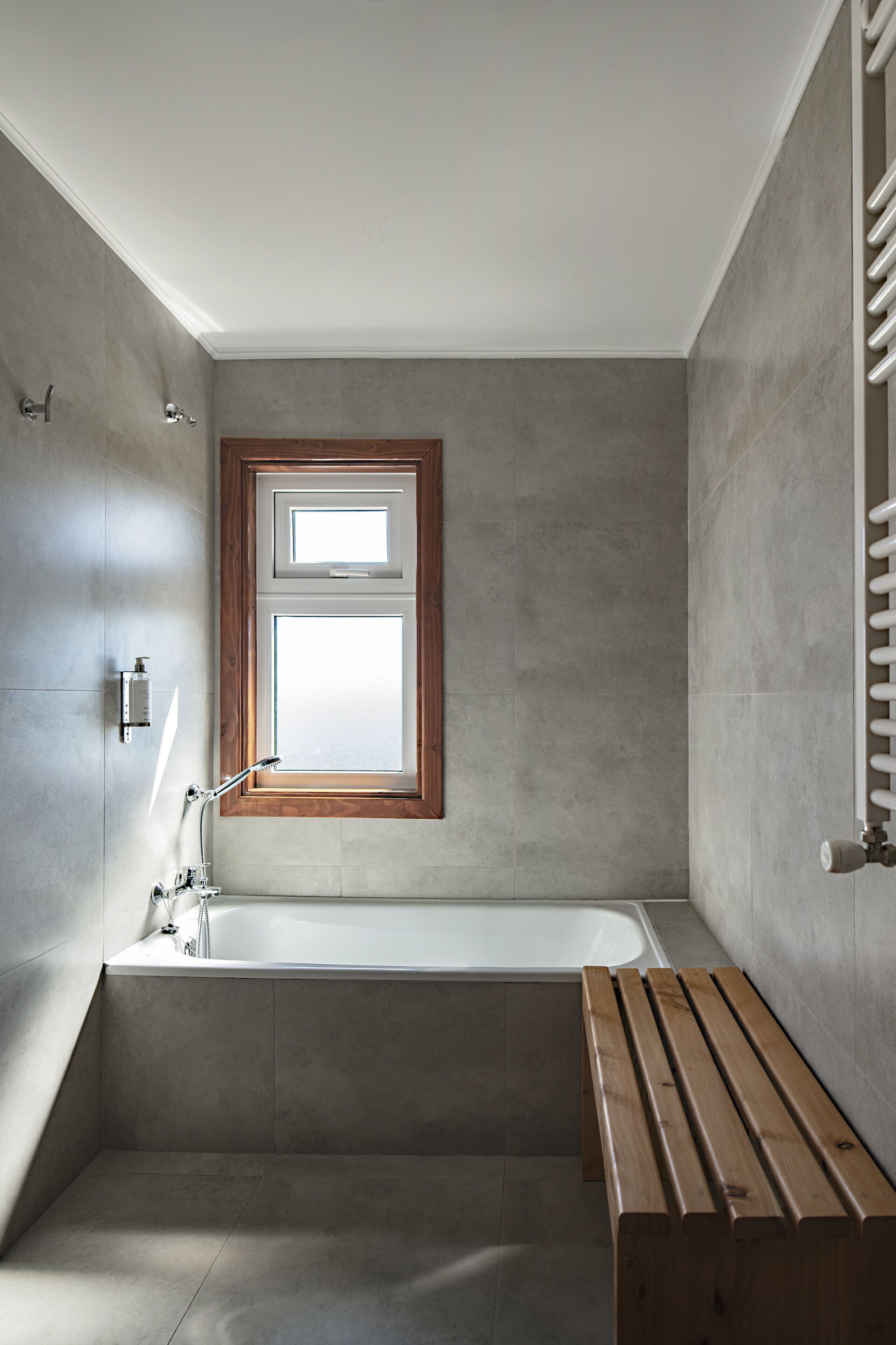 A modern bathroom with a freestanding bathtub, a wooden bench, and a window with a wooden frame.