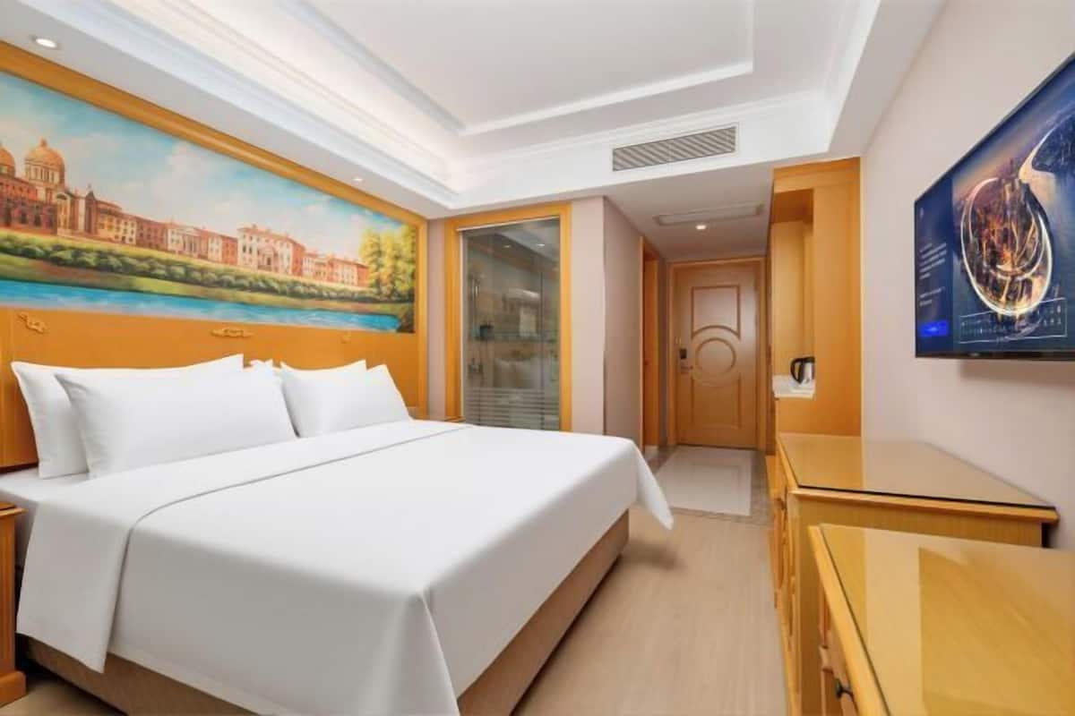 Una habitación de hotel con una cama, un escritorio, un cuadro de un paisaje urbano y un televisor montado en la pared.